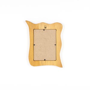 May include: A light brown wooden picture frame with a wavy, organic shape. The back of the frame is visible, showing a cardboard insert and a small metal hanger. The frame has a natural wood grain texture.