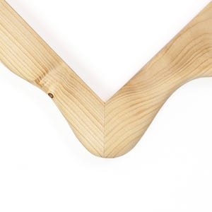 Puede incluir: Una percha de madera de color claro con un dise&ntilde;o en forma de V. La veta natural de la madera es claramente visible en toda la superficie, mostrando la textura org&aacute;nica y los tonos bronceados claros del material en la uni&oacute;n central.