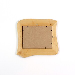 May include: A rectangular picture frame made of light-coloured natural wood with an irregular, wavy outer edge. The back of the frame features a brown fibreboard backing secured with small metal clips and two metal hanging hooks attached.