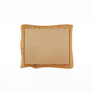 May include: A rectangular picture frame with a natural light brown wood finish and an irregular, wavy outer edge. The back of the frame features a flat, tan-colored backing board secured with metal clips and two metal hanging brackets attached.