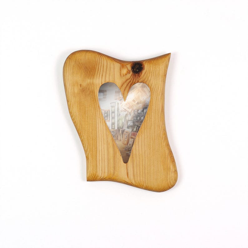 Puede incluir: Un marco de fotos de madera con un recorte en forma de coraz&oacute;n. El marco es de madera clara con vetas visibles y un nudo. La abertura en forma de coraz&oacute;n revela una fotograf&iacute;a. El marco tiene una forma ondulada y org&aacute;nica.