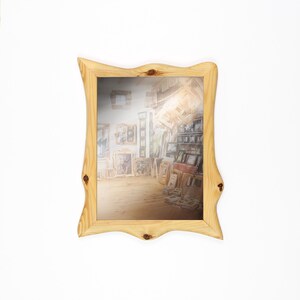 May include: A rectangular mirror with a light-colored, natural wood frame. The frame has a unique, wavy shape with knots. The mirror reflects a room filled with artwork and frames, creating a gallery-like appearance. The mirror is set against a white background.