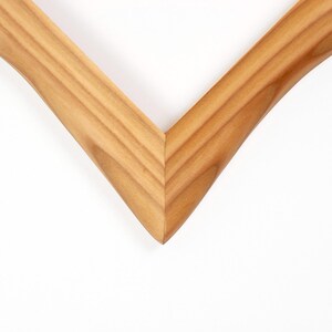 K&ouml;nnte beinhalten: Nahaufnahme einer Ecke eines Holzbilderrahmens. Der Rahmen besteht aus hellbraunem Holz mit sichtbarer Maserung und einem V-f&ouml;rmigen Design. Der Rahmen befindet sich vor einem wei&szlig;en Hintergrund.