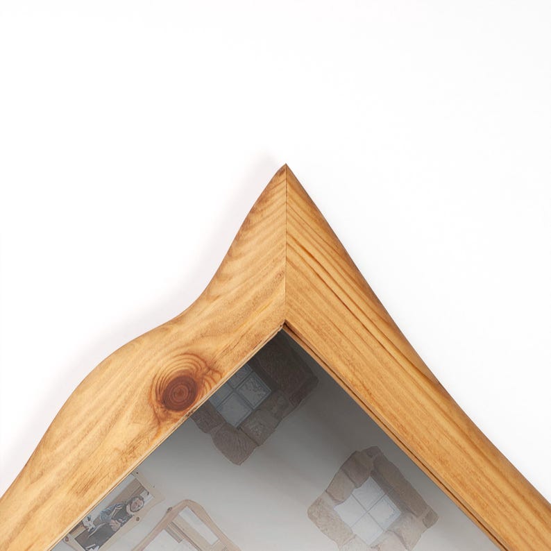 May include: A wooden picture frame with a natural finish. The frame has a unique, pointed top and a curved side. The glass reflects a blurred image of a room. The frame is made of light-coloured wood.