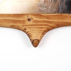 Pu&ograve; includere: Primo piano di una cornice in legno con finitura naturale venata. La cornice ha un design curvo unico con un dettaglio a nodo nella parte inferiore. La cornice &egrave; in legno marrone chiaro.