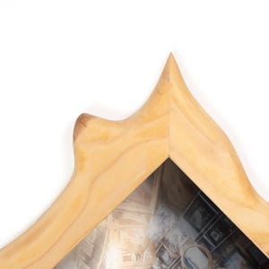 Peut inclure: Un cadre en bois clair avec une forme unique en forme de montagne. Il poss&egrave;de une vitre transparente r&eacute;v&eacute;lant un collage d'images et un petit miroir rond. Le grain du bois est visible, ajoutant &agrave; l'esth&eacute;tique naturelle.