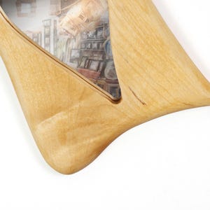 May include: A light-colored wooden picture frame with a smooth, organic, and curved shape. The frame features a triangular cutout that holds a photograph displaying an interior scene with various framed pictures and a woven basket on a shelf.