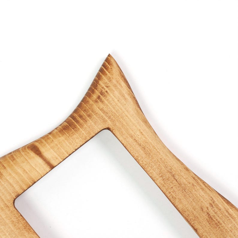 May include: Close-up of a wooden clothes hanger with a distinctive geometric shape. The hanger is crafted from light-coloured wood, showcasing the grain and a natural finish. It has a pointed top and a rectangular opening.