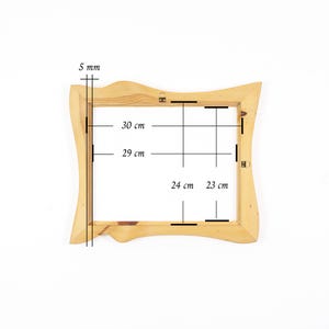 May include: A light-coloured wooden picture frame with an irregular, wavy outer edge. Technical lines and text indicate dimensions of 5 mm, 30 cm, 29 cm, 24 cm, and 23 cm. The frame features two small metal hanging brackets attached to the top and side.