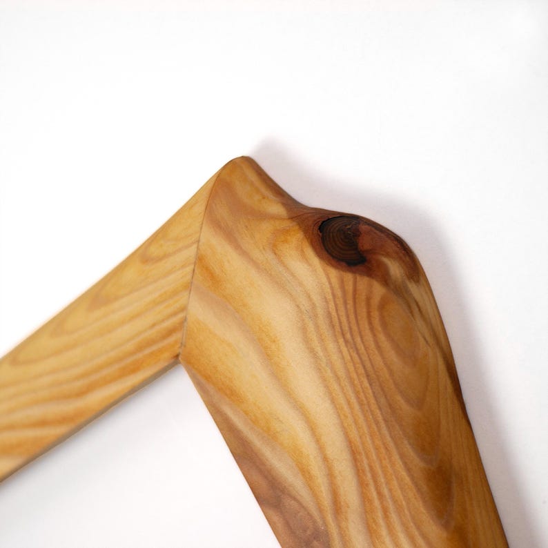 Puede incluir: Una secci&oacute;n de esquina de un marco de madera con un patr&oacute;n de veta de madera natural de color marr&oacute;n claro. La superficie muestra un detalle de nudo circular oscuro prominente cerca de la uni&oacute;n a inglete donde dos piezas de madera se unen.