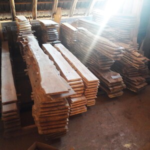 Puede incluir: Apilados de tablas de madera sin terminar en un entorno r&uacute;stico. Las tablas var&iacute;an en tama&ntilde;o y forma, algunas con bordes naturales. La madera es de color marr&oacute;n claro con vetas visibles. Las tablas est&aacute;n apiladas en montones ordenados, listas para proyectos de carpinter&iacute;a.