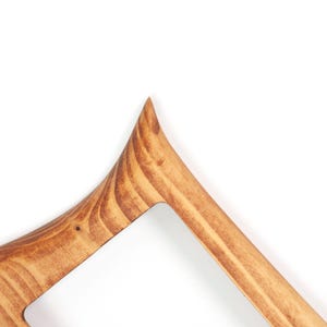May include: Close-up of a wooden picture frame corner. The frame is made of light brown wood with visible grain patterns. The design features a pointed top and a rectangular opening.