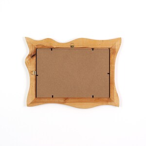 May include: A rectangular picture frame crafted from light-toned natural wood with an irregular, wavy outer edge. The back of the frame features a flat brown backing board secured with metal clips and includes two metal hanging hooks attached.