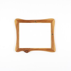 Op de afbeelding: Een houten fotolijst met een natuurlijke afwerking. Het frame heeft een uniek, gebogen ontwerp met een knoopdetail aan de onderrand. Inclusief ophangmateriaal aan de achterkant. Het frame is leeg, klaar voor een foto of kunstwerk.