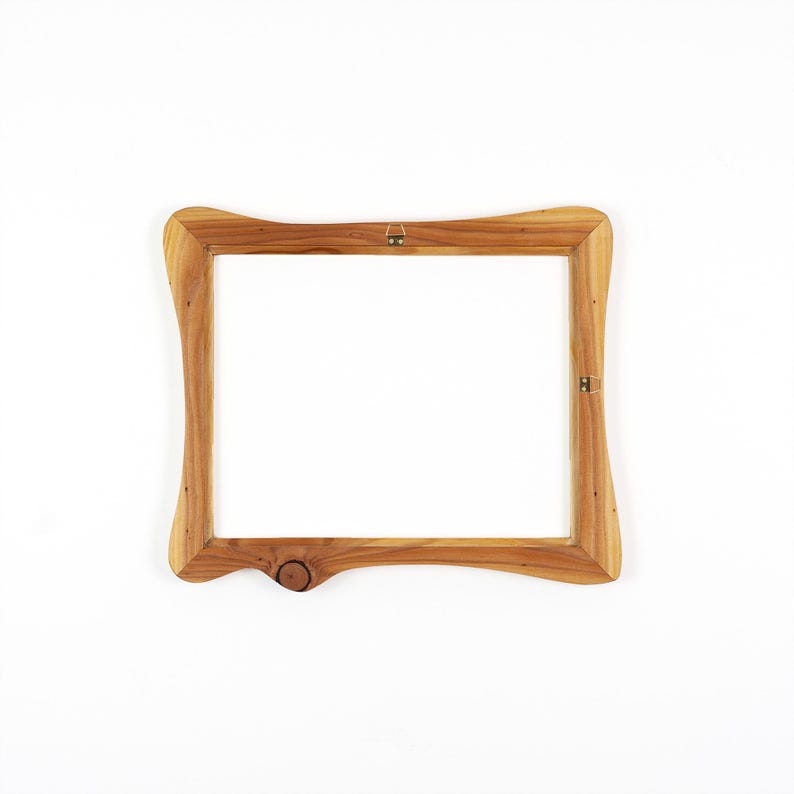 Op de afbeelding: Een houten fotolijst met een natuurlijke afwerking. Het frame heeft een uniek, gebogen ontwerp met een knoopdetail aan de onderrand. Inclusief ophangmateriaal aan de achterkant. Het frame is leeg, klaar voor een foto of kunstwerk.