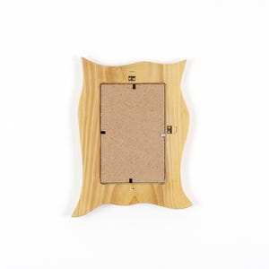 May include: A rectangular wooden picture frame with a wavy, organic outer edge. The frame is made of light-colored natural wood with visible grain patterns. The back of the frame features a brown fiberboard backing with metal hanging hardware.