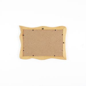 May include: A rectangular wooden picture frame shown from the back, featuring a light natural wood finish with wavy, organic edges. The centre is filled with a flat, brown fibreboard backing secured by small metal clips and a metal hanging bracket.