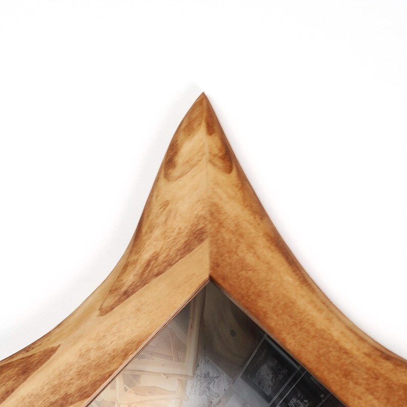 May include: Close-up of a wooden frame with a unique, pointed top. The frame has a light brown color with visible wood grain. The frame appears to be holding a picture or artwork, with a glimpse of the contents visible through the glass.