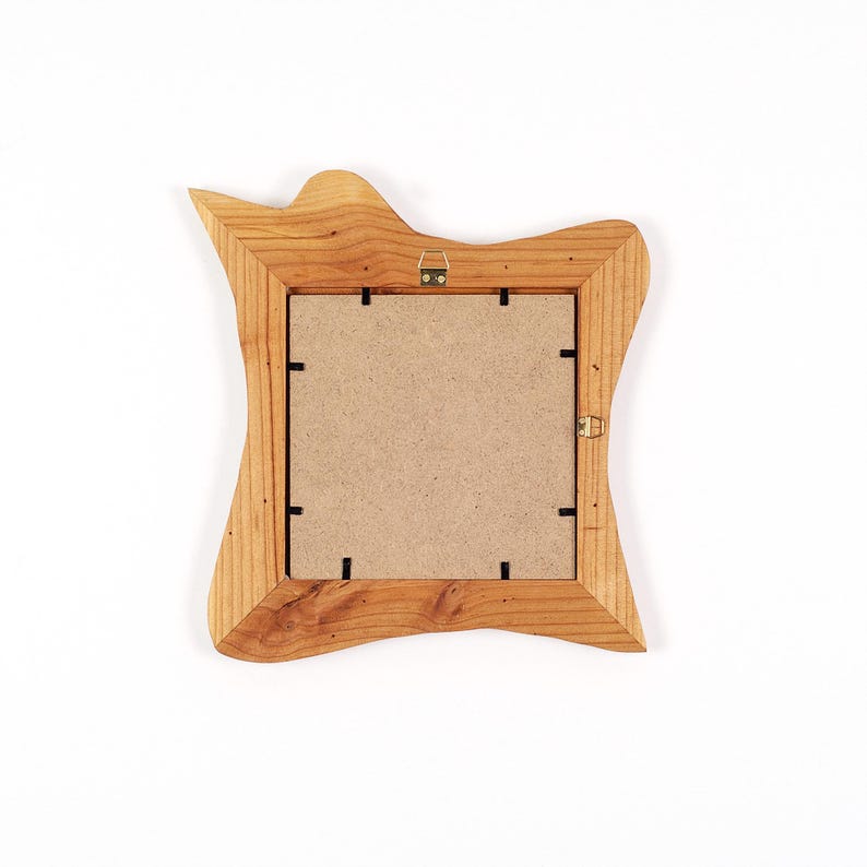 Pu&ograve; includere: Una cornice quadrata con una finitura naturale in venature di legno marrone chiaro e un bordo esterno irregolare e organico. Il retro della cornice presenta un pannello in fibra beige fissato con clip metalliche nere e due ganci dorati.