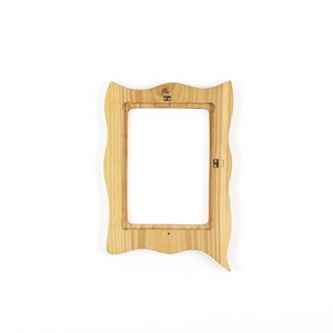 May include: A light-colored wooden picture frame with a wavy, irregular border. The frame has a rectangular opening and hardware for hanging. The wood grain is visible, and the frame is set against a white background.