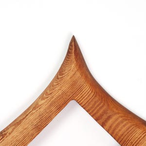 May include: A close-up shot of a wooden clothes hanger with a distinctive, angular shape. The hanger is crafted from light brown wood, highlighting the natural grain pattern. The top of the hanger is pointed, giving it a contemporary feel.