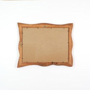 May include: A wooden picture frame with a wavy edge design. The frame is made of a light brown wood and has a cardboard backing. The frame has a brass hanging hook and a brass clasp.