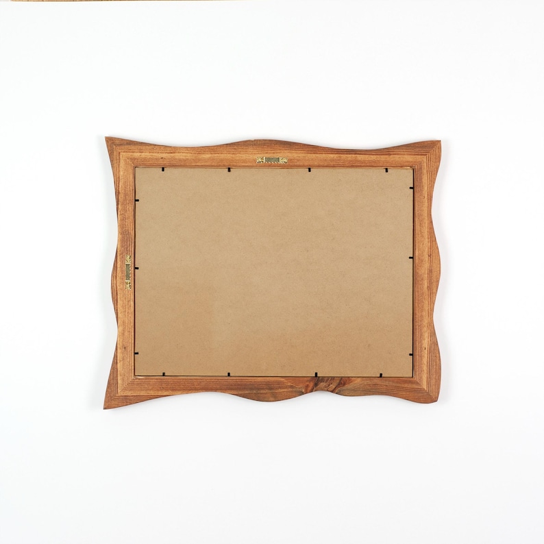 May include: A wooden picture frame with a wavy edge design. The frame is made of a light brown wood and has a cardboard backing. The frame has a brass hanging hook and a brass clasp.