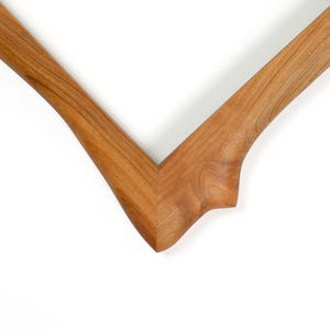 Puede incluir: Una esquina de marco de fotos de madera en forma de V con un patr&oacute;n de veta de madera marr&oacute;n claro natural. El marco tiene un acabado liso y pulido con una curva org&aacute;nica distintiva en el borde exterior donde las dos piezas de madera se unen.