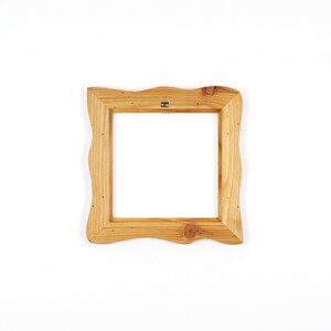 Puede incluir: Un marco de fotos cuadrado de madera con un acabado natural en marr&oacute;n claro y un borde exterior ondulado y org&aacute;nico. El marco presenta un patr&oacute;n de veta de madera visible y un peque&ntilde;o soporte met&aacute;lico para colgar en el centro superior.