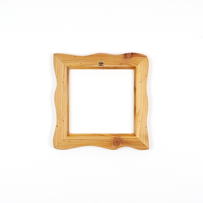 Puede incluir: Un marco de fotos cuadrado de madera con un acabado natural en marr&oacute;n claro y un borde exterior ondulado y org&aacute;nico. El marco presenta un patr&oacute;n de veta de madera visible y un peque&ntilde;o soporte met&aacute;lico para colgar en el centro superior.