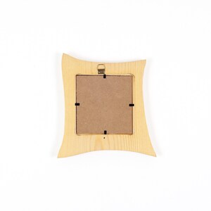 May include: A square wooden picture frame with a light tan natural wood grain finish and curved side edges. The back of the frame features a brown fibreboard backing secured by four black metal clips and a small gold metal hanging bracket at the top.