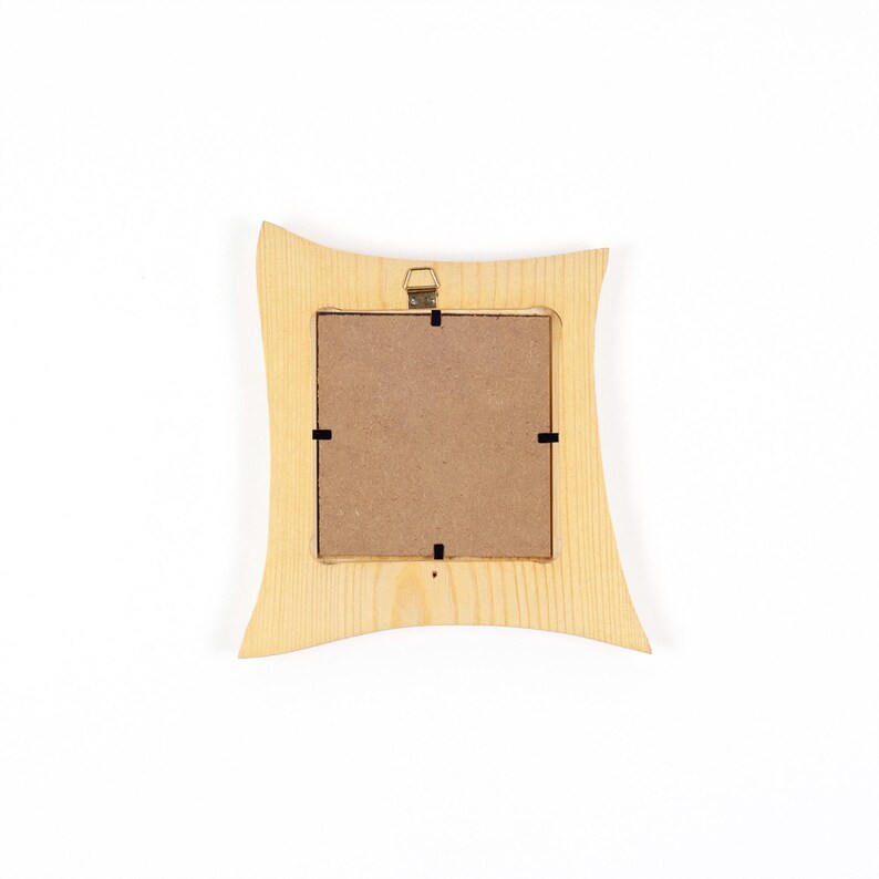May include: A square wooden picture frame with a light tan natural wood grain finish and curved side edges. The back of the frame features a brown fibreboard backing secured by four black metal clips and a small gold metal hanging bracket at the top.