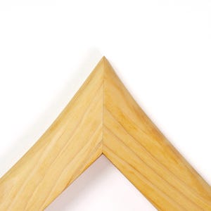 Puede incluir: Una secci&oacute;n de esquina de un marco de fotos de madera de color claro con una uni&oacute;n a inglete. La madera muestra un patr&oacute;n de veta natural con vetas de color marr&oacute;n claro visibles y una superficie lisa e inclinada que se une en una punta.