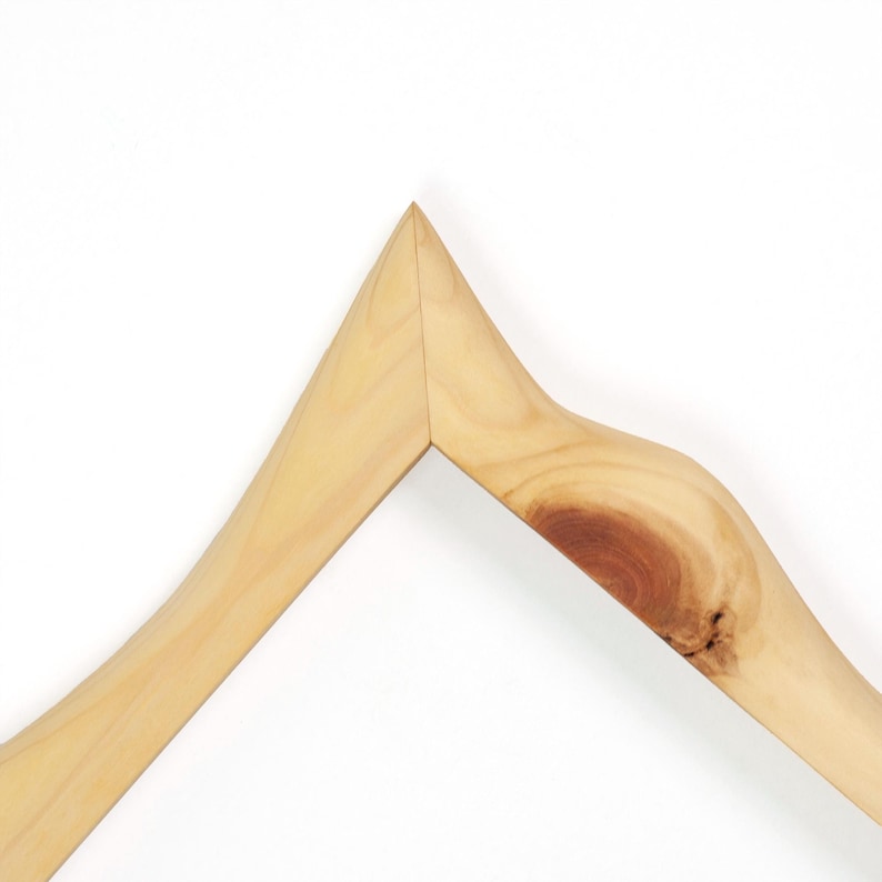 May include: A light-coloured wooden clothes hanger featuring a prominent natural wood grain pattern and a distinct dark brown knot detail on the right side. The hanger has a sharp, angular peak at the top centre and smooth, curved edges throughout.