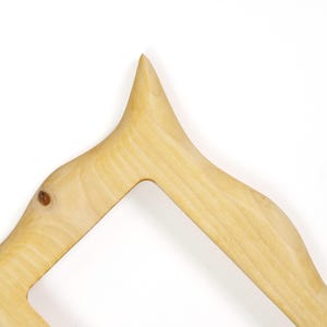 Pu&ograve; includere: Una cornice in legno di colore chiaro con una finitura liscia e naturale e venature visibili. La cornice presenta un angolo superiore appuntito e bordi curvi, che mettono in risalto la consistenza organica e le tonalit&agrave; marrone chiaro del legno.