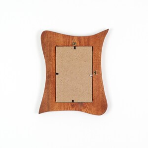 May include: Rear view of a wooden picture frame with a distinctive, undulating shape. The frame is a warm brown hue, featuring a rectangular aperture for a photograph. The back has a cardboard backing and metal hanging hardware.