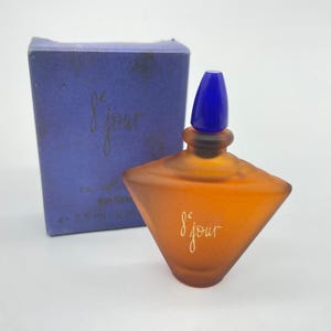 Puede incluir: Un frasco de perfume de color naranja con tapón azul y caja a juego. El frasco tiene una forma geométrica única y las palabras "8 jour" están escritas en oro en el frasco y la caja. La caja es de color azul oscuro.