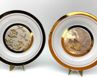 Könnte beinhalten: Zwei weiße Keramikteller mit goldenen Rändern und einem erhabenen, geprägten Design aus Vögeln und Blumen. Die Teller stehen auf goldenen Metallständern.