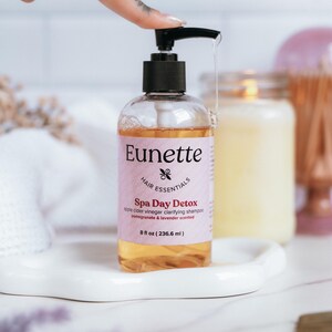 May include: A clear bottle of apple cider vinegar clarifying shampoo with a pink label that says "Eunette Hair Essentials Spa Day Detox pomegranate & lavender scented 8 fl oz (236.6 ml)"