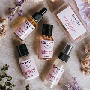 May include: Four bottles of hair care products with the brand name "Eunette Hair Essentials" and the product names "Spa Day Feels", "Spa Day Replenish", "Spa Day Detox", and "Spa Day Refresh". The bottles are arranged on a light gray surface with flowers and greenery.
