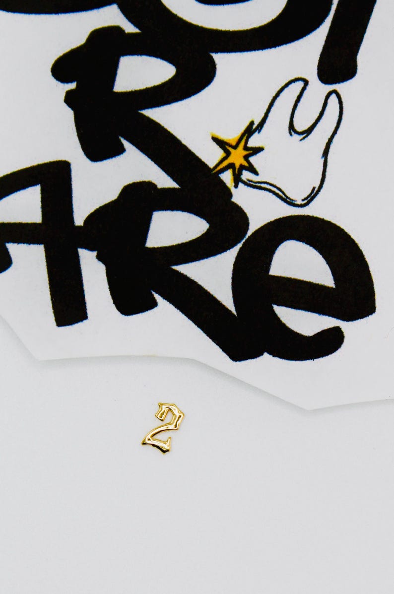 May include: Gold-colored number two charm with a gothic font style. The charm is on a white background.