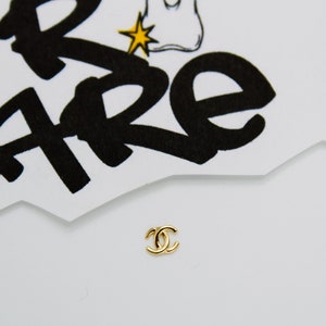 May include: A gold-colored Chanel logo charm. The logo is a double 'C' with a rounded shape.