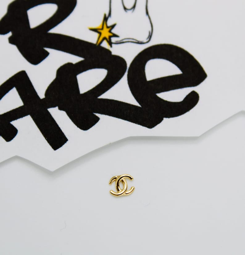 May include: A gold-colored Chanel logo charm. The logo is a double 'C' with a rounded shape.