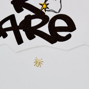 May include: A gold-colored spider charm on a white background. The charm is small and detailed, with eight legs and a body.