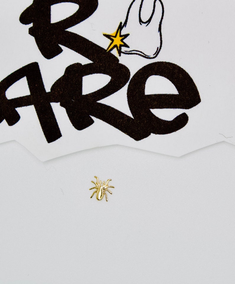 May include: A gold-colored spider charm on a white background. The charm is small and detailed, with eight legs and a body.