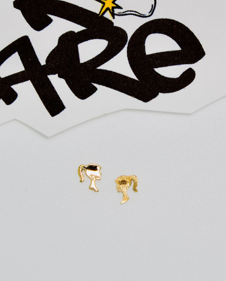 May include: A pair of gold-colored earrings shaped like a woman's profile with a ponytail.
