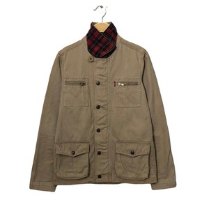 May include: A tan jacket with a red and black plaid collar. The jacket has two chest pockets with zippers, two lower flap pockets, and a button-up front. A small red tag is visible on the chest.