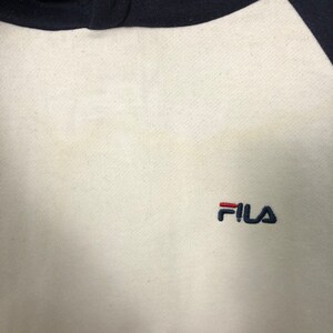 May include: A cream-colored sweatshirt featuring dark navy blue raglan sleeves and a dark navy blue collar. The brand name FILA is embroidered on the chest in dark navy blue thread, with the letter F featuring a horizontal red bar accent.