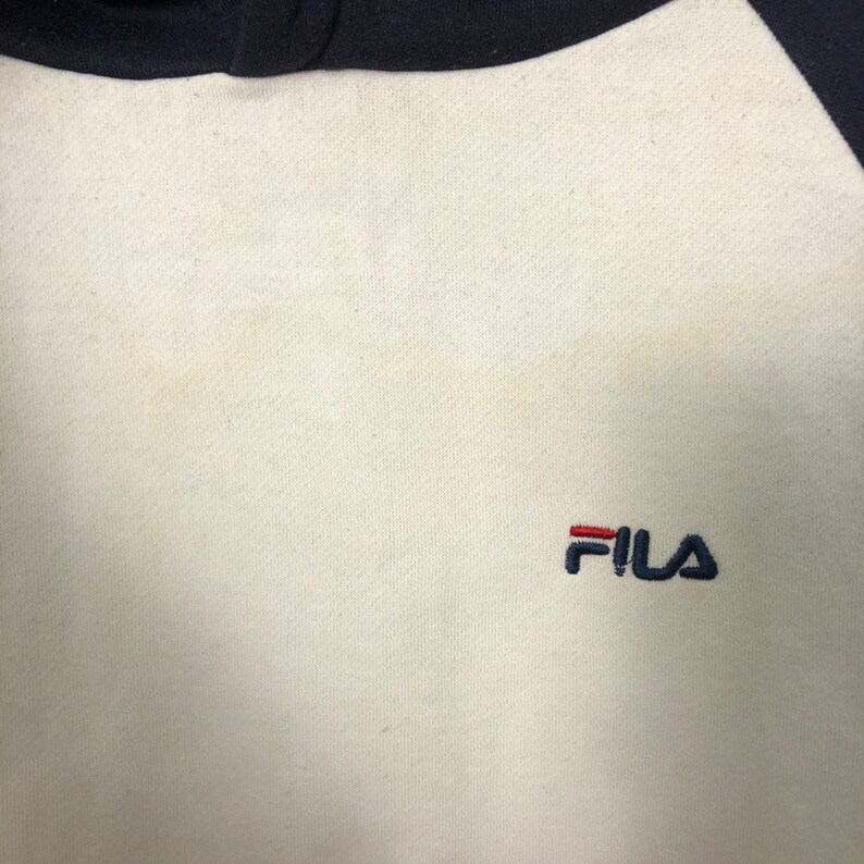 May include: A cream-colored sweatshirt featuring dark navy blue raglan sleeves and a dark navy blue collar. The brand name FILA is embroidered on the chest in dark navy blue thread, with the letter F featuring a horizontal red bar accent.