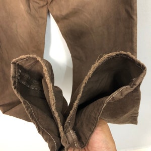 May include: Close-up of brown denim pants. The pants are a solid color with a slightly faded appearance. The cuffs are turned up, revealing the raw edge of the fabric. The pants appear to be well-worn.